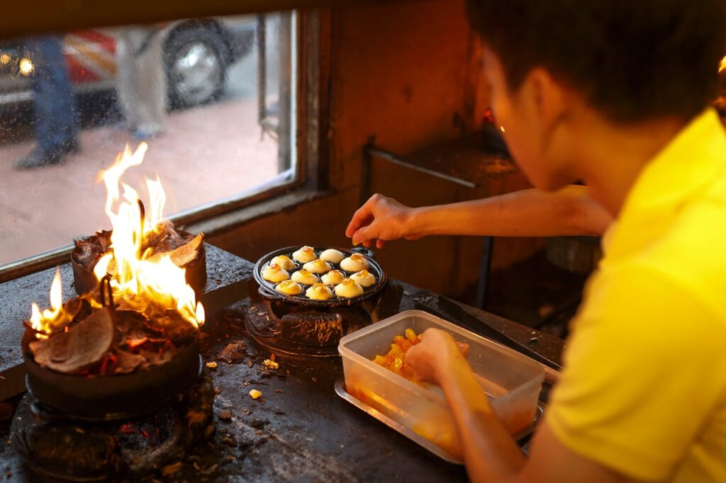 A street vendor prepares a traditional dessert over open flames in an indoor stall.